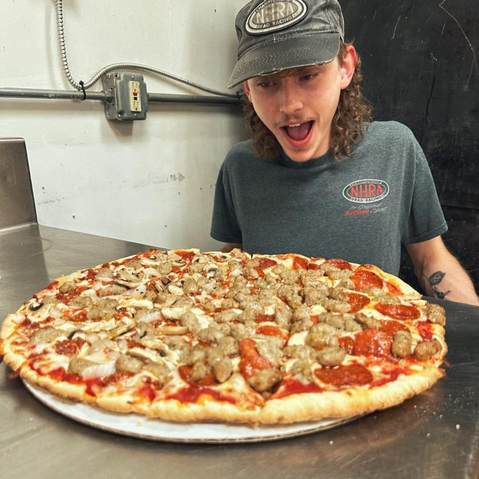A U-Know Pizza employee excitedly looks at a large pizza with pepperoni, sausage, and mushrooms.