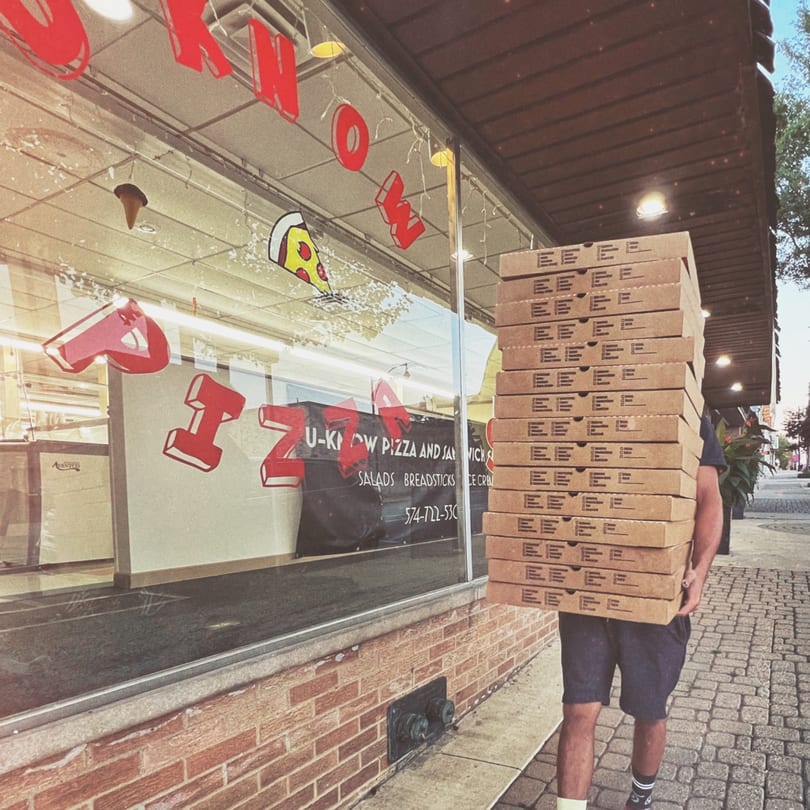 Person carrying a tall stack of U-Know Pizza boxes outside the restaurant's storefront.