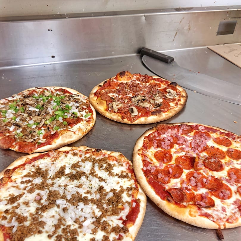 Four U-Know Pizza & Sandwich Shop pizzas with various toppings on a metal counter.