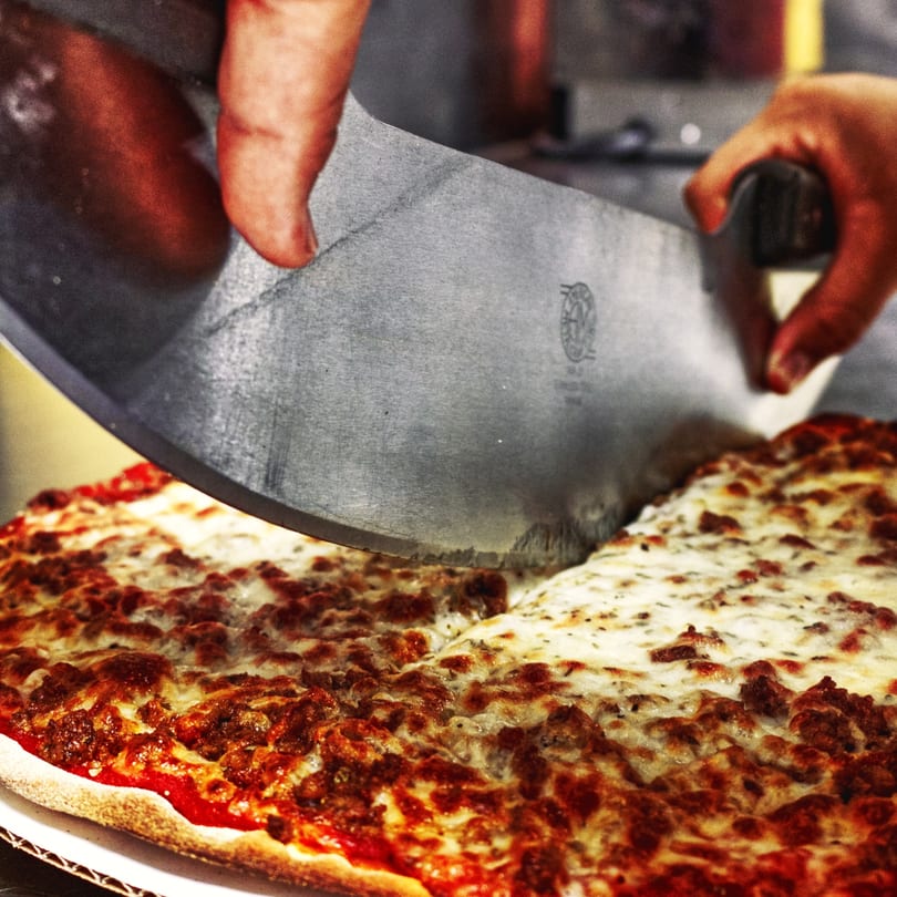 Hands using a large pizza cutter to slice a freshly baked U-Know Pizza.